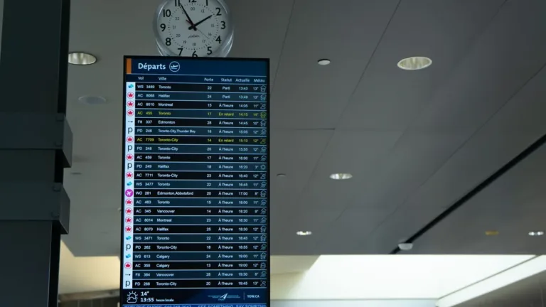 Airport departures screen listing flight times and statuses, with a wall clock mounted above it in a terminal