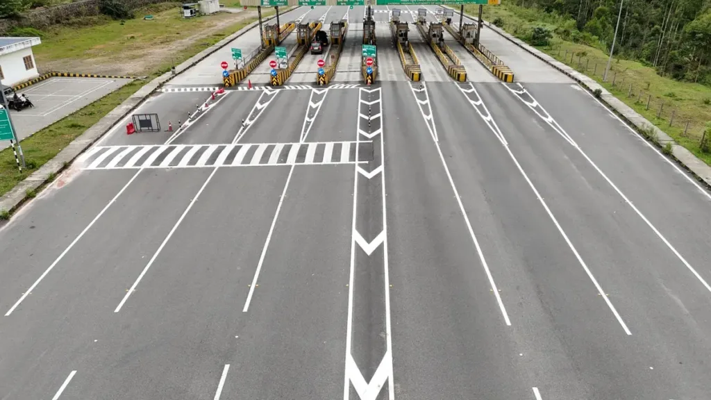 Aerial view of a wide multi-lane highway leading to a toll plaza canopy with booths, set among green hills