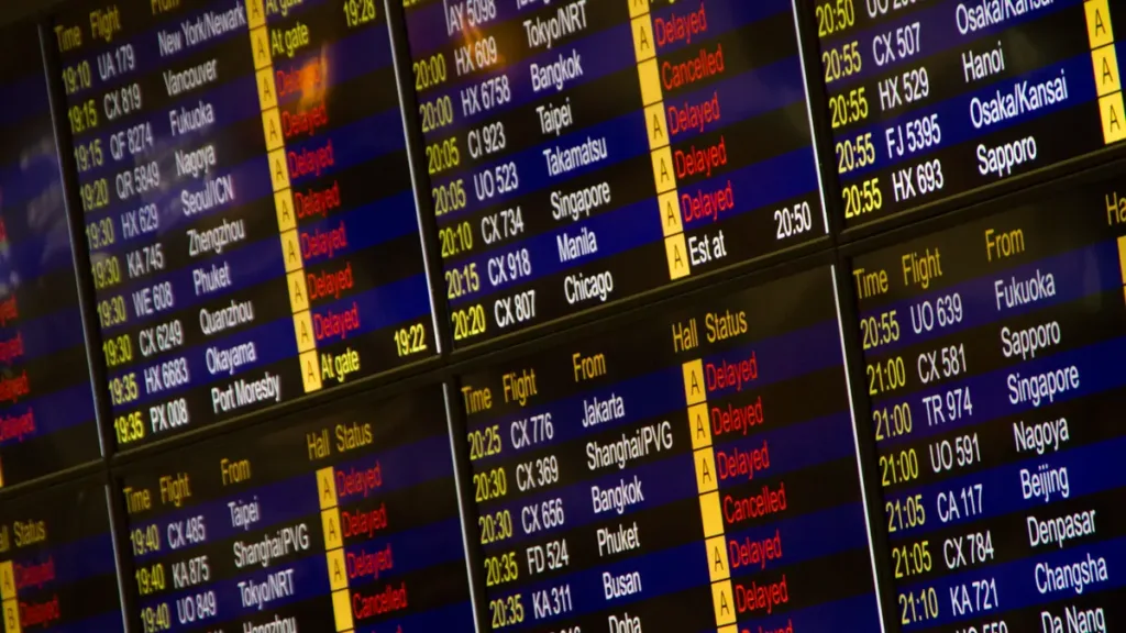 Close-up of an airport flight information screen listing flight times, origins and status lines such as Delayed