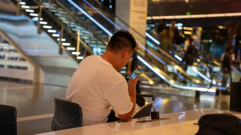 Traveller seen from behind using a smartphone at a counter in a modern airport terminal, escalators behind