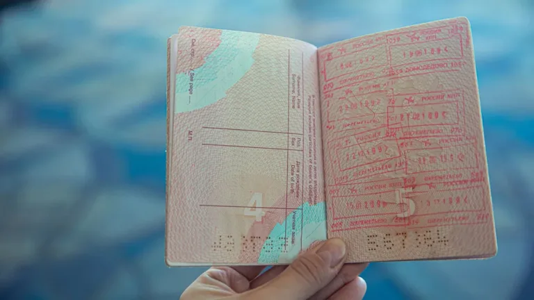Hand holding an open passport showing pages of entry and exit stamps against a blurred blue airport background