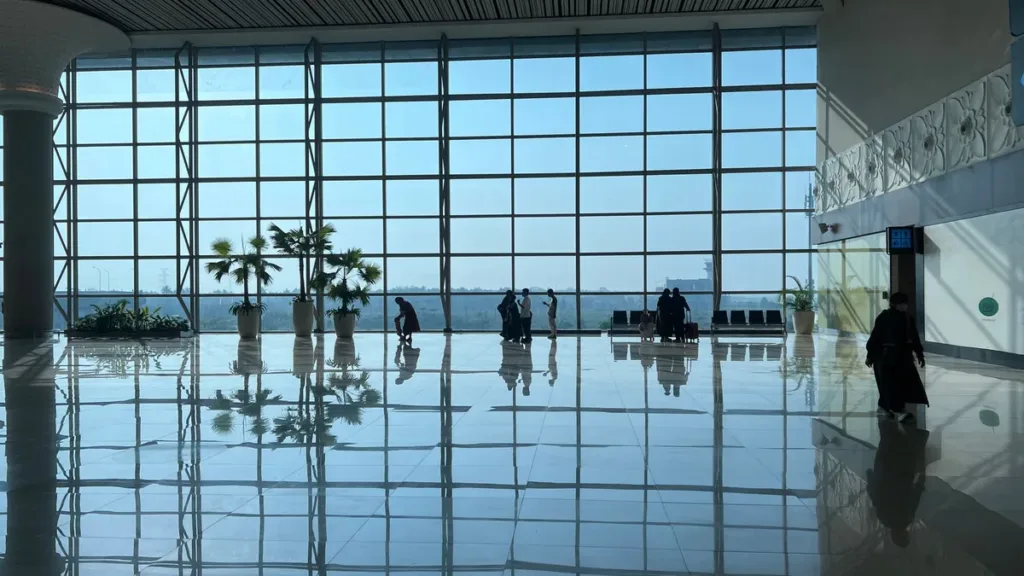 Bright airport terminal hall with floor-to-ceiling windows, a polished floor and a few travellers in silhouette