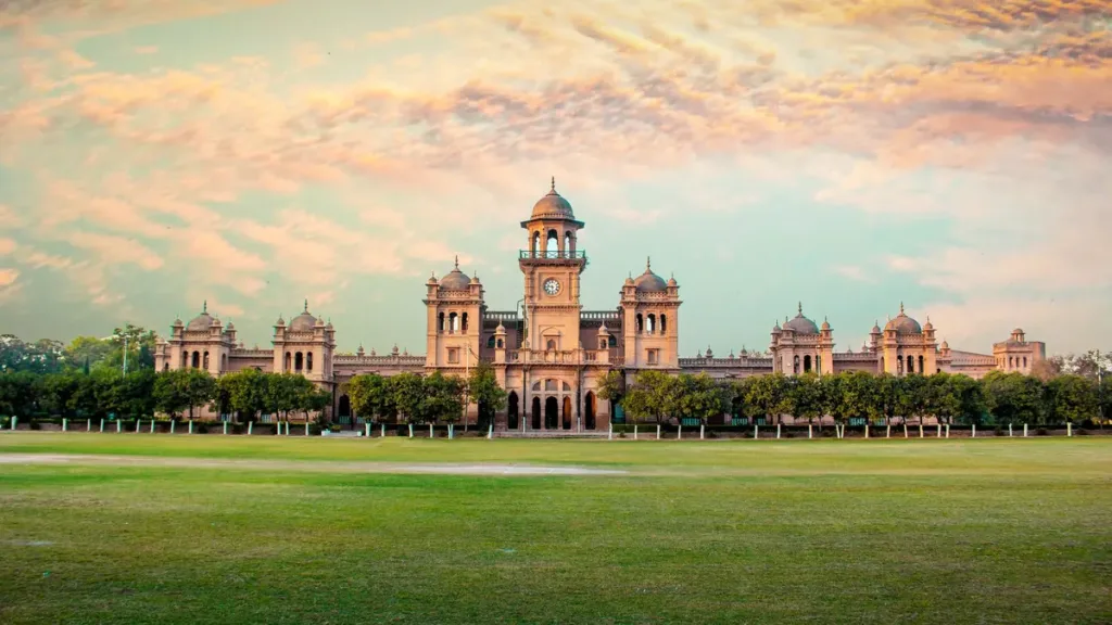 Islamia College Peshawar's red-brick clock tower and domes behind a wide green lawn under a pink evening sky
