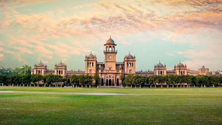 Islamia College Peshawar's red-brick clock tower and domes behind a wide green lawn under a pink evening sky