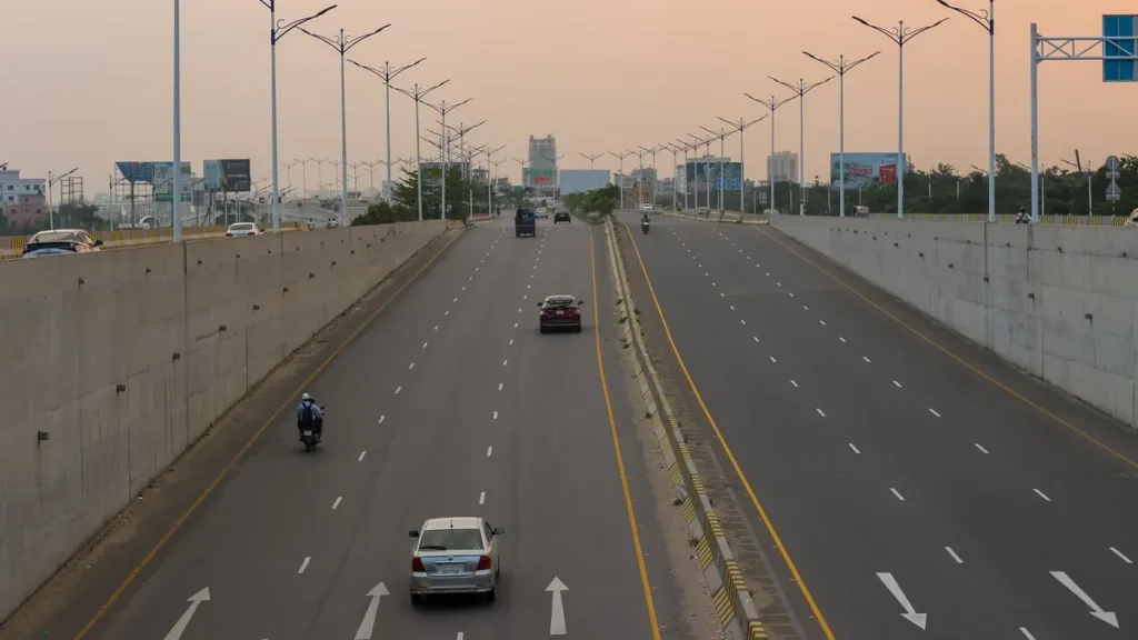 Wide divided city road at dusk with light traffic, a car and a motorcyclist, painted lane arrows and rows of streetlights