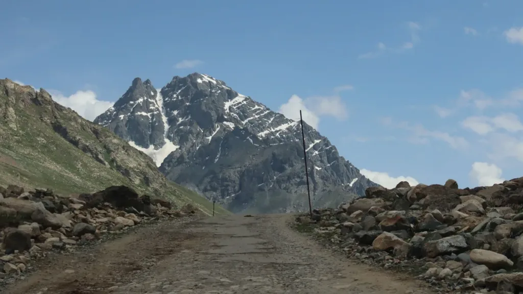 Unpaved gravel mountain road lined with rocks leading toward jagged snow-capped peaks under a blue sky