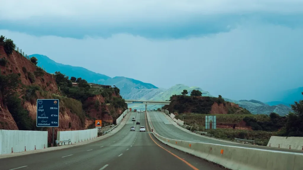 Divided motorway in Pakistan curving past hills under a cloudy sky, with a blue direction sign and an overpass ahead
