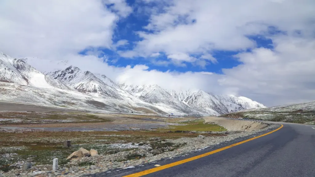 Empty mountain highway curving across a high plateau towards snow-capped peaks under a cloudy blue sky