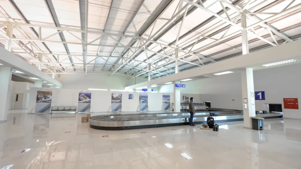 Quiet, brightly lit airport baggage reclaim hall with an empty carousel under a white steel roof and a lone traveller waiting