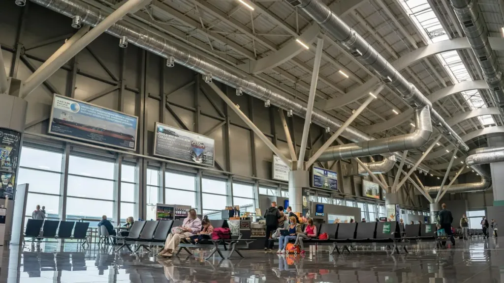 Passengers seated and walking in a bright airport terminal hall with a steel roof and tall glass walls