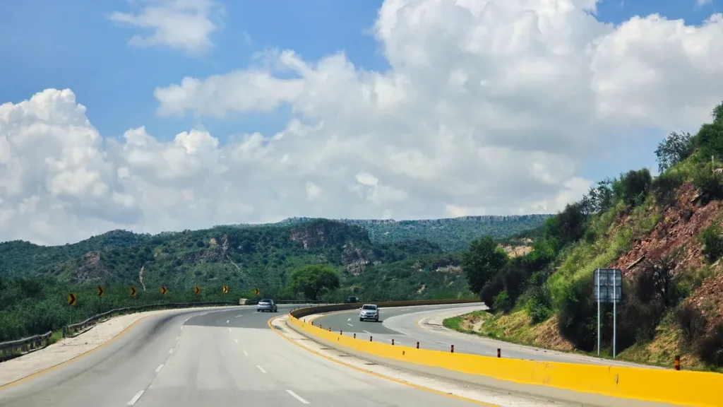 A curving dual carriageway with yellow kerbs winding through green wooded hills under a bright sky with white clouds