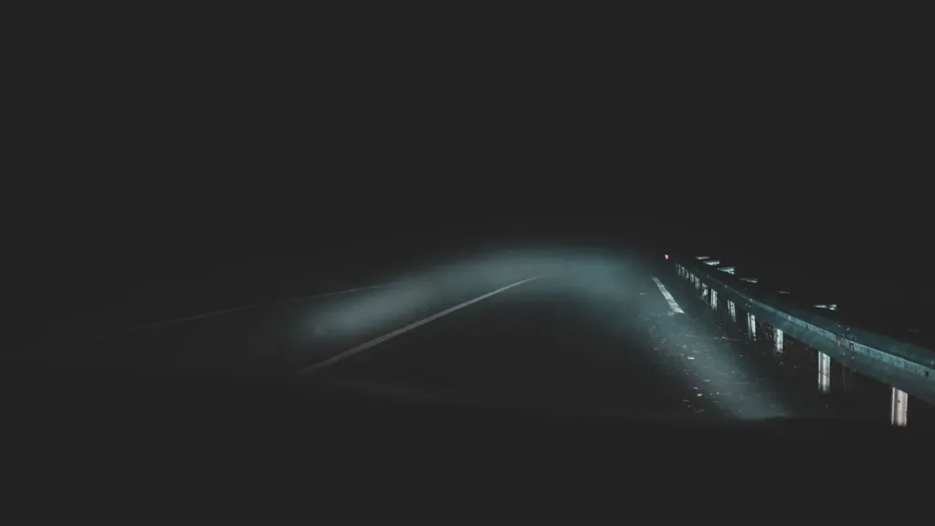 Dark road at night swallowed by thick fog, headlight beams lighting a short stretch of tarmac beside a metal guardrail