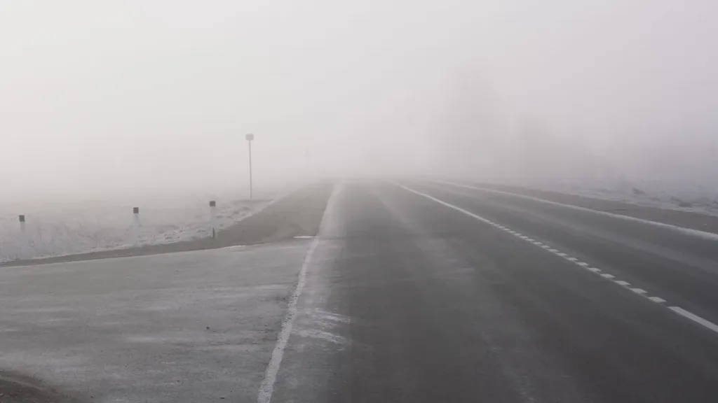 Empty multi-lane highway disappearing into dense fog, with lane markings and roadside posts fading from view