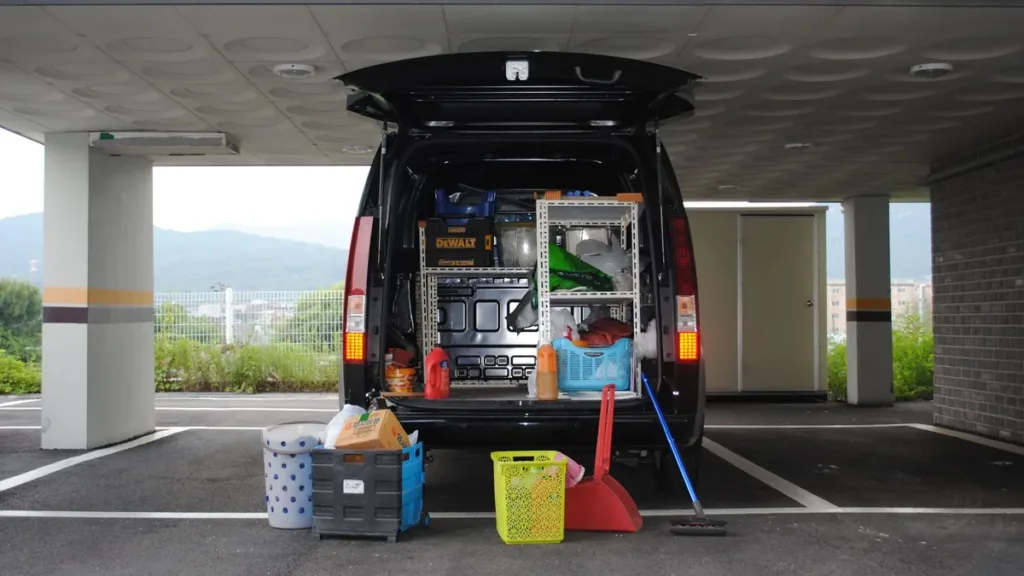 Rear view of a dark van with its tailgate open, shelves and equipment loaded inside, parked under a covered bay