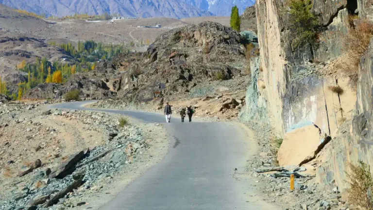 Narrow paved road winding past a rock face through a dry mountain valley, with two distant walkers and autumn trees
