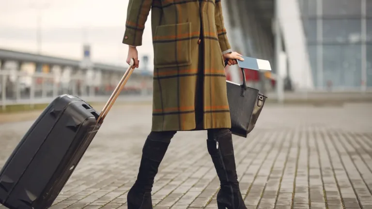 Business traveller in a long coat pulling a black wheeled suitcase and carrying a bag and papers outside an airport terminal