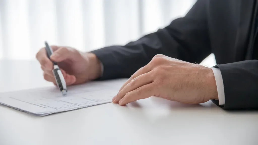 Hand of a person in a dark suit signing a printed document with a pen at a white desk