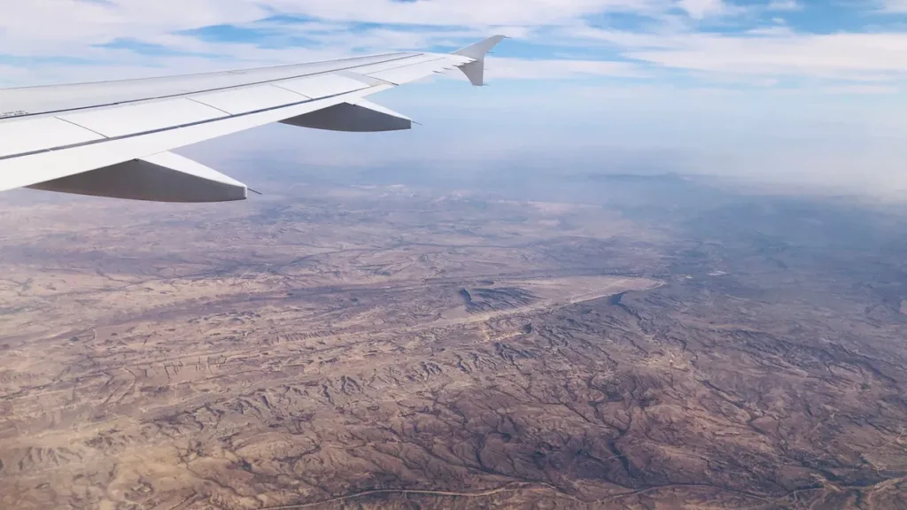 Airliner wing above a vast, dry desert landscape of ridges and plains, seen from a passenger window under hazy sky