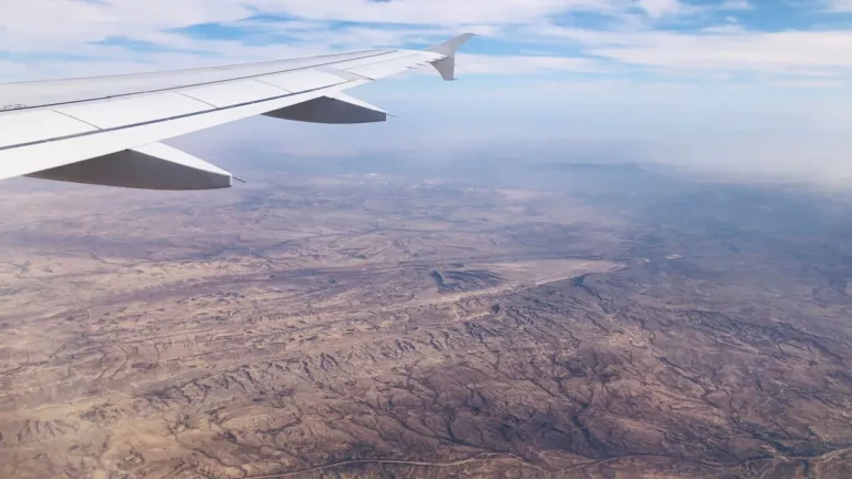 Airliner wing above a vast, dry desert landscape of ridges and plains, seen from a passenger window under hazy sky