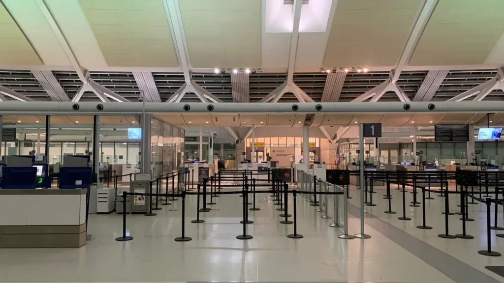 Quiet airport check-in hall with empty queue lanes, rope barriers and counters under a bright white ceiling