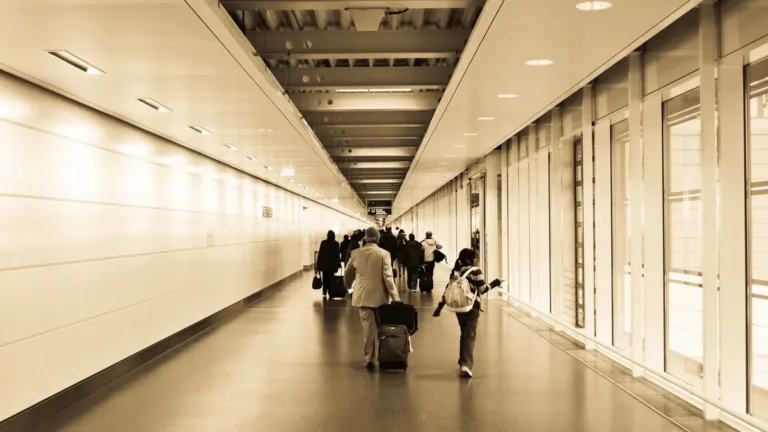 Travellers wheeling suitcases down a long, bright airport walkway towards the terminal, in warm sepia tones