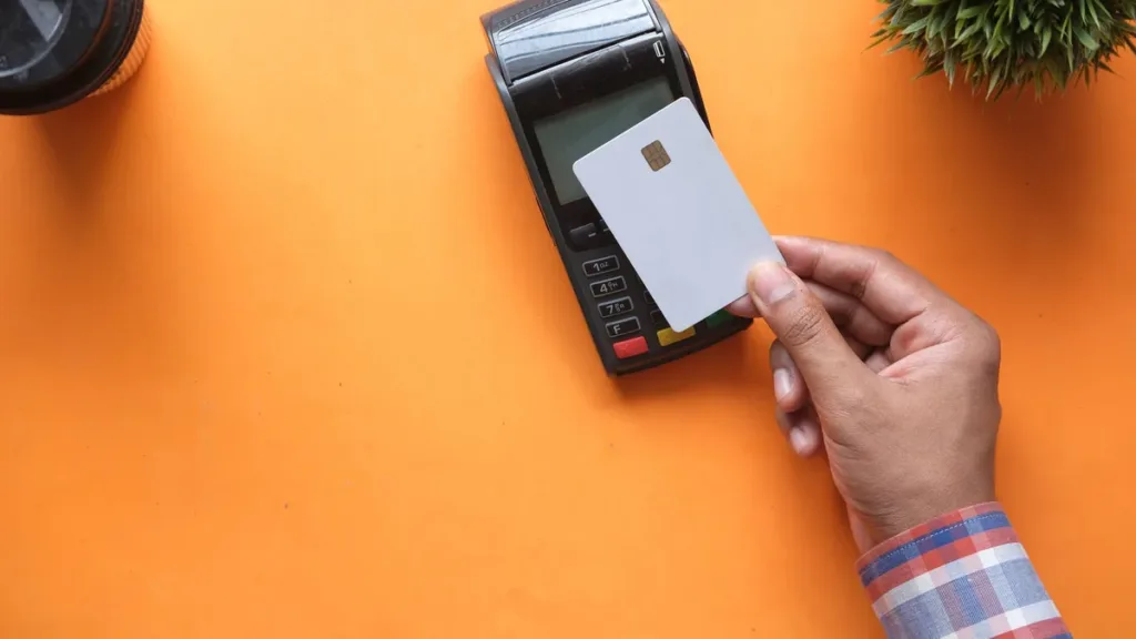 Hand holding a plain white bank card against a black card payment terminal on an orange tabletop