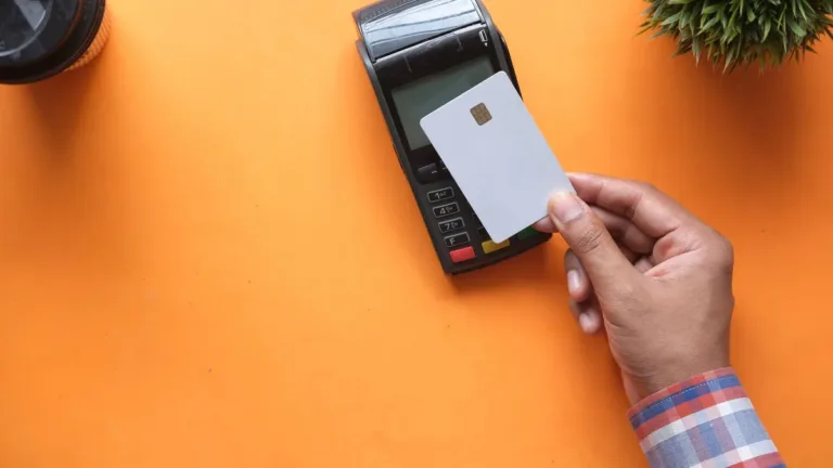 Hand holding a plain white bank card against a black card payment terminal on an orange tabletop