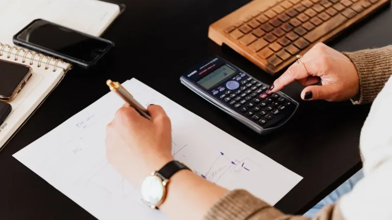 Hands working out costs with a calculator and pen on paper at a dark desk, beside a phone and a wooden keyboard