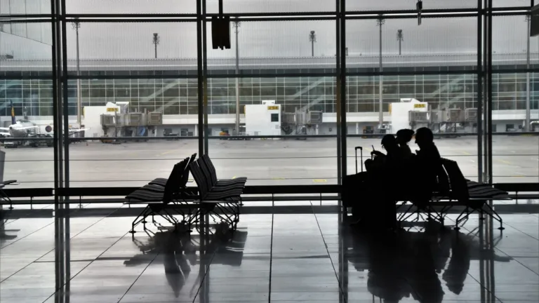 Silhouetted travellers waiting on seats with luggage beside tall windows of an airport terminal