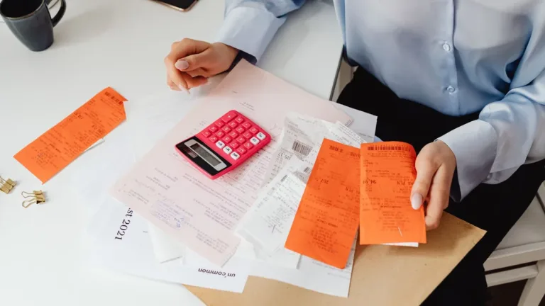Hands sorting orange and white paper receipts beside a pink calculator on a desk while reviewing expenses