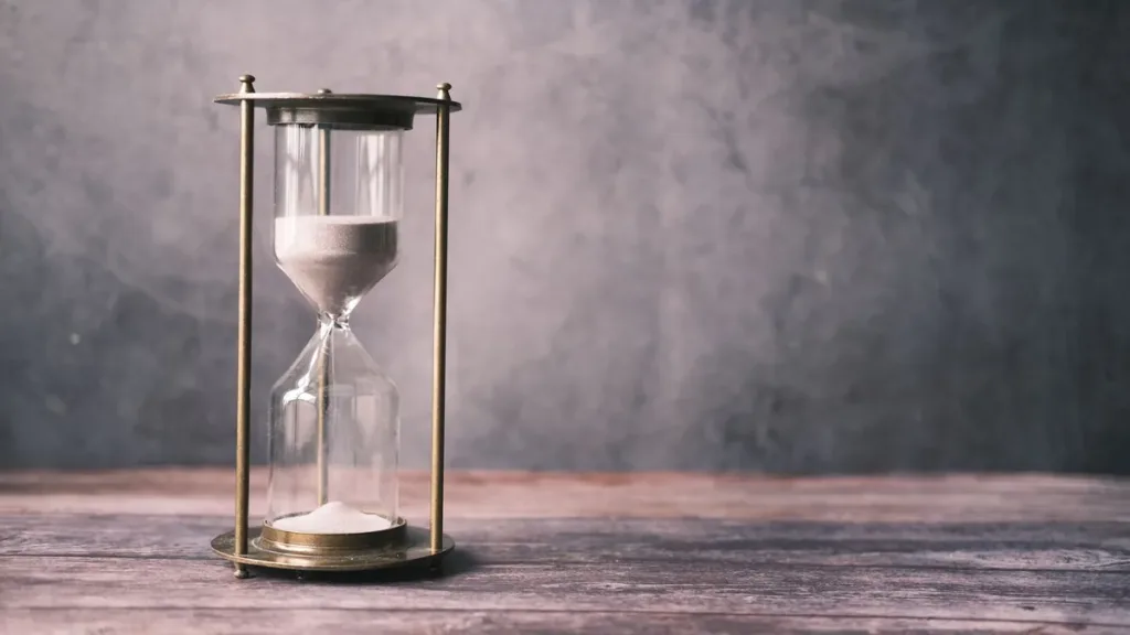 Brass-framed hourglass with white sand running down, standing on a weathered wooden table against a grey wall