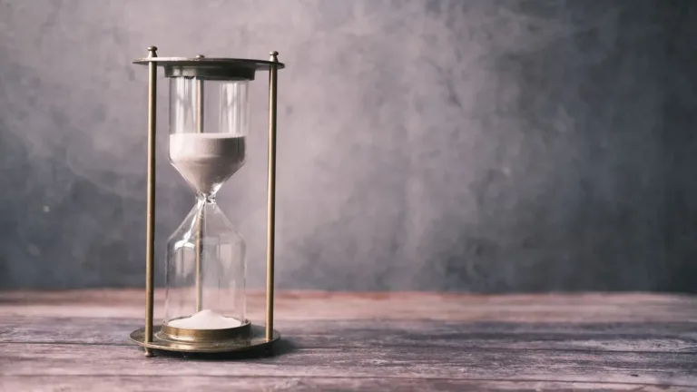 Brass-framed hourglass with white sand running down, standing on a weathered wooden table against a grey wall