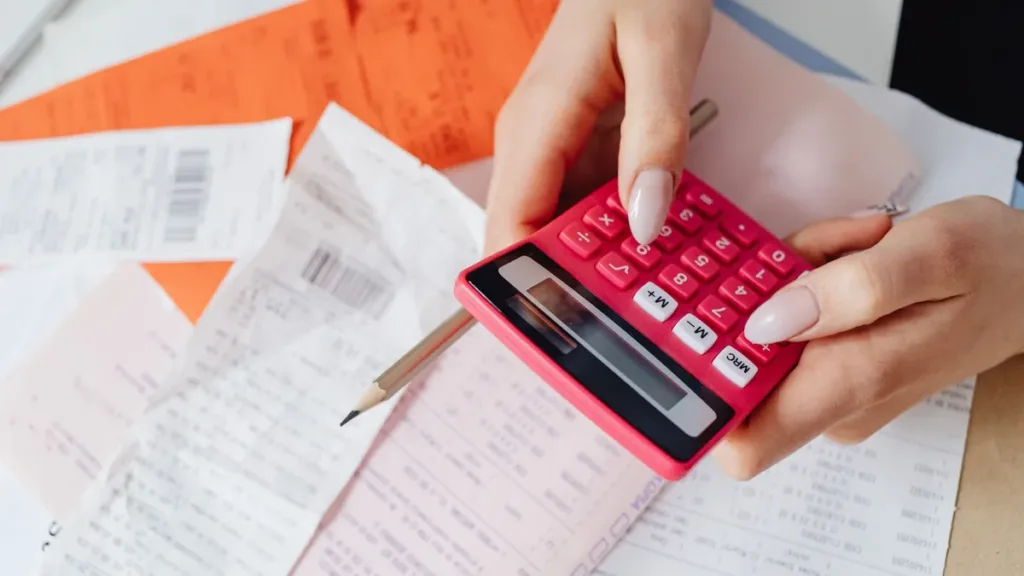Hands tallying expenses on a pink calculator over a spread of printed receipts and invoices on a desk