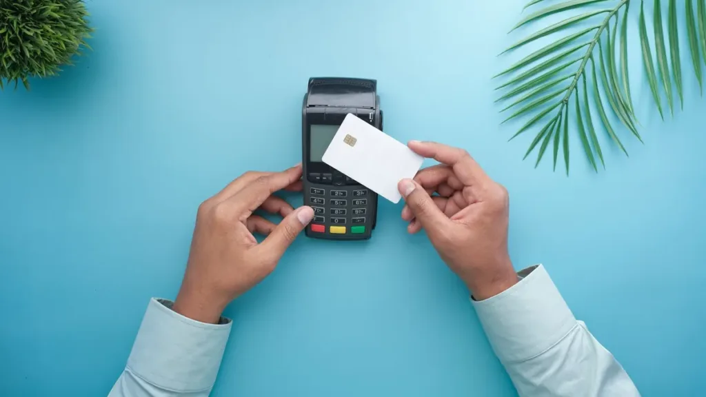 Two hands holding a plain white bank card beside a card payment terminal on a light blue background with palm leaves