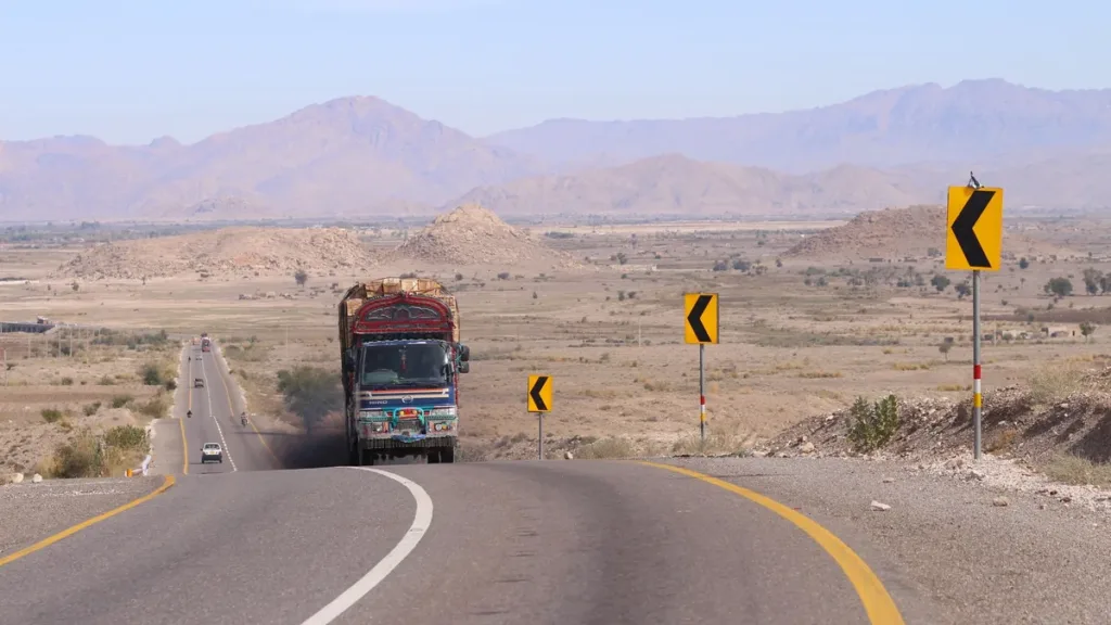 Decorated truck on a long curving highway through arid plains and mountains near Khuzdar, with yellow chevron signs
