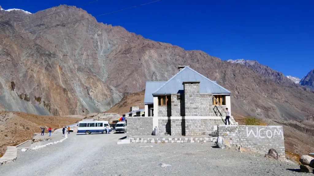 Stone rest house on a mountain roadside with a white van and cars parked on the gravel below bare rocky peaks