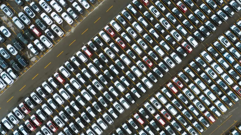 Aerial view of rows of mostly white and grey cars parked tightly on a large vehicle lot
