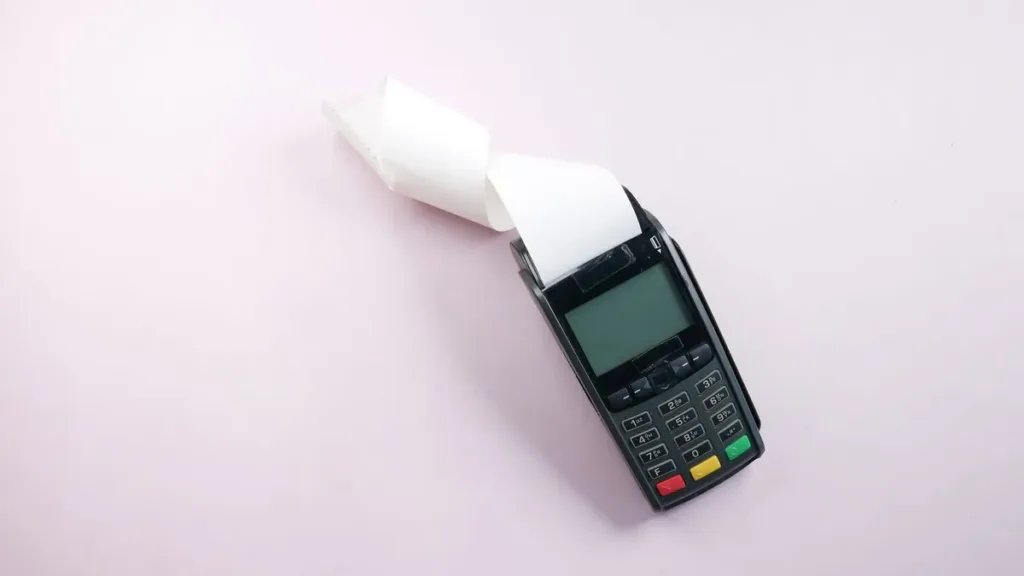 A black card payment terminal printing a curled paper receipt, shot from above on a plain light pink surface
