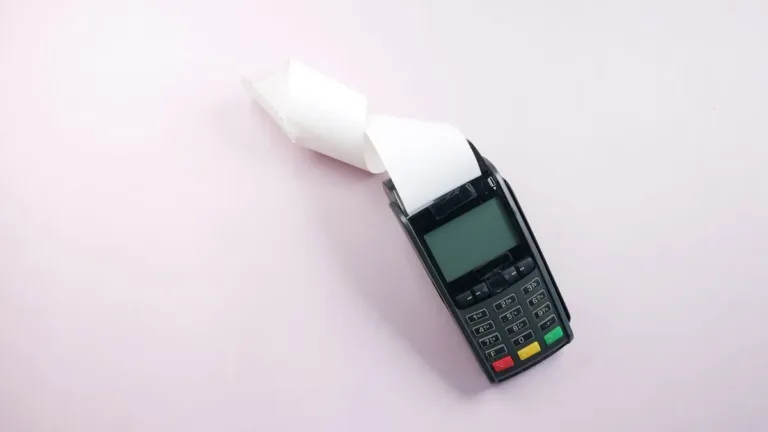 A black card payment terminal printing a curled paper receipt, shot from above on a plain light pink surface