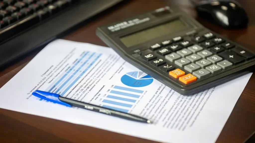 Black desk calculator and blue pen resting on a printed financial report with bar and pie charts beside a keyboard