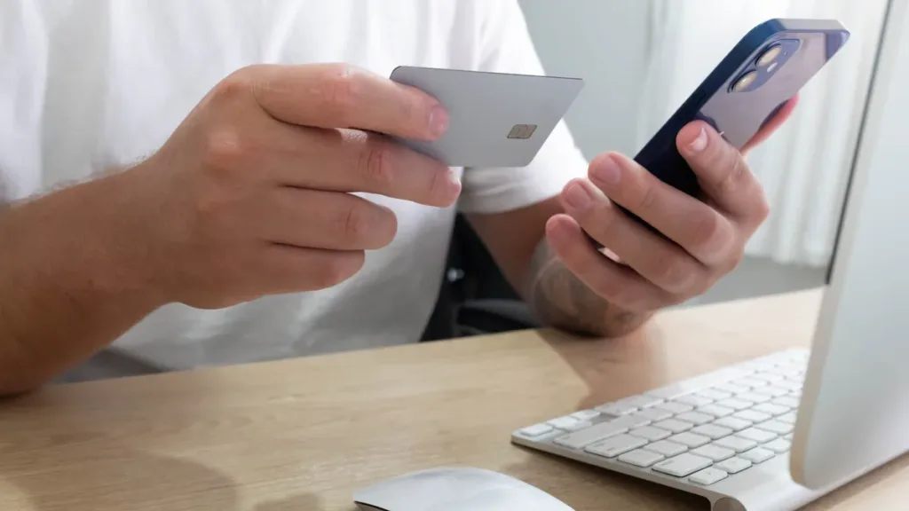 Hands holding a blank bank card and a smartphone at a desk beside a computer keyboard and mouse