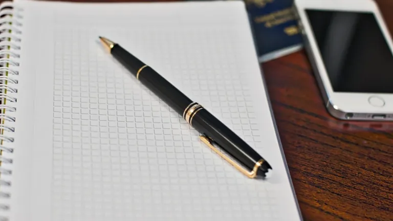 Grid notebook with a black pen beside a smartphone and a passport partly visible on a wooden desk