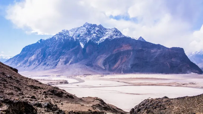 Snow-capped mountain rising above a wide sandy river plain and rocky slopes in the Skardu valley under a blue sky