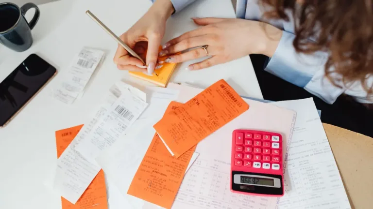 Hands sorting printed receipts beside a pink calculator and paperwork on a desk, preparing an expense claim