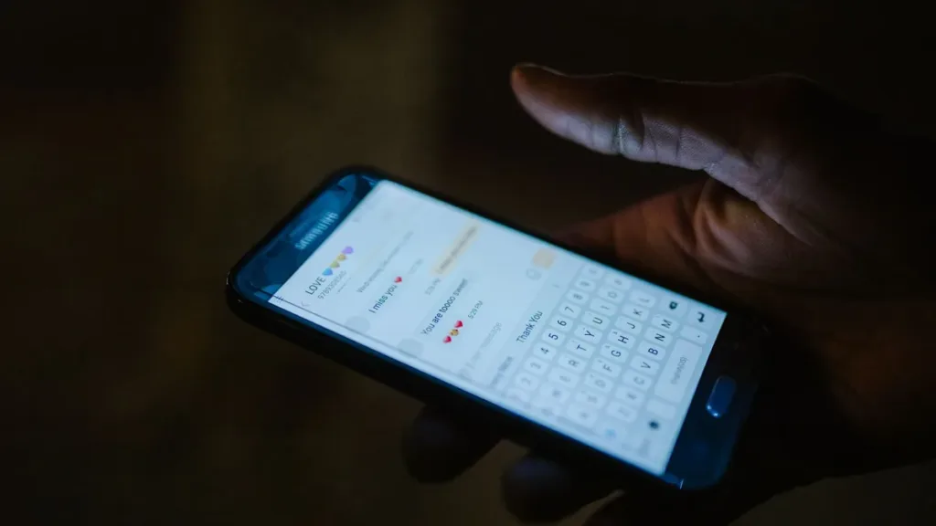 Close-up of a hand holding a smartphone with a messaging app and keyboard open, lit by the screen in a dark room