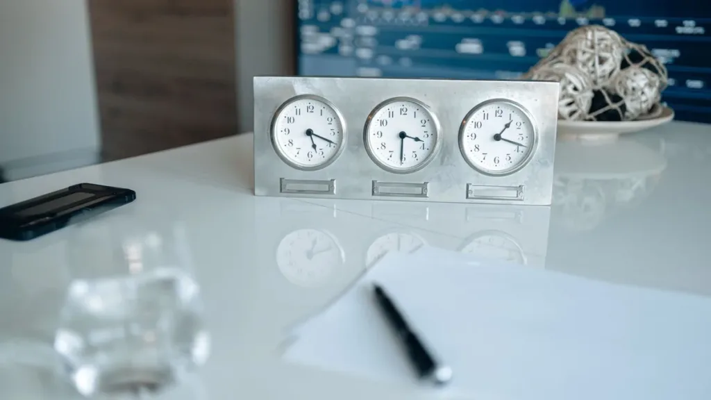 Three silver desk clocks showing different time zones on a white desk beside a pen and notepad