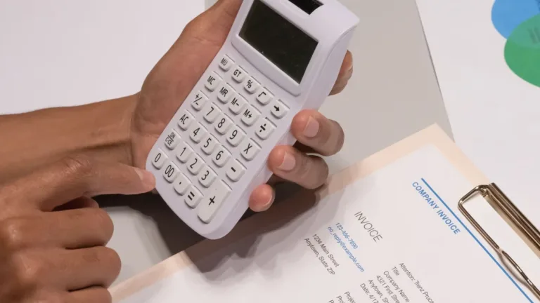 Hands holding a calculator over a printed invoice on a clipboard, checking figures line by line