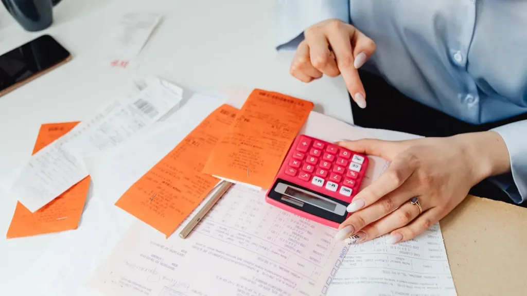 Hands checking a stack of paper receipts and invoices with a pink calculator on an office desk