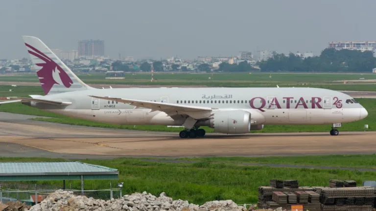 A Qatar Airways Boeing 787 Dreamliner taxiing along a runway beside green grass, with city buildings in the distance