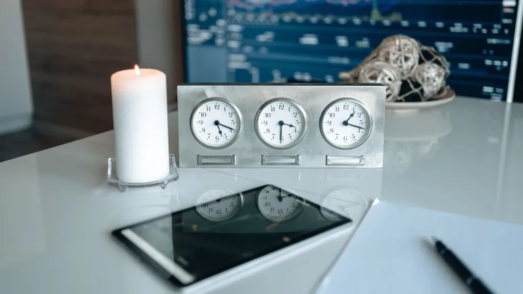 Three small analogue desk clocks set to different times beside a candle and a tablet on a glossy white office desk